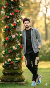 A young man leaning casually against a flower-covered tree in a park during golden hour, wearing a black outfit and sneakers, with a blurred natural background.