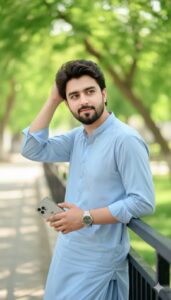 The image shows a young man standing outdoors in a green park, casually leaning on a railing while fixing his hair with one hand and holding a phone in the other. He looks relaxed and confident in a light blue outfit, with soft natural light and a blurred leafy background.